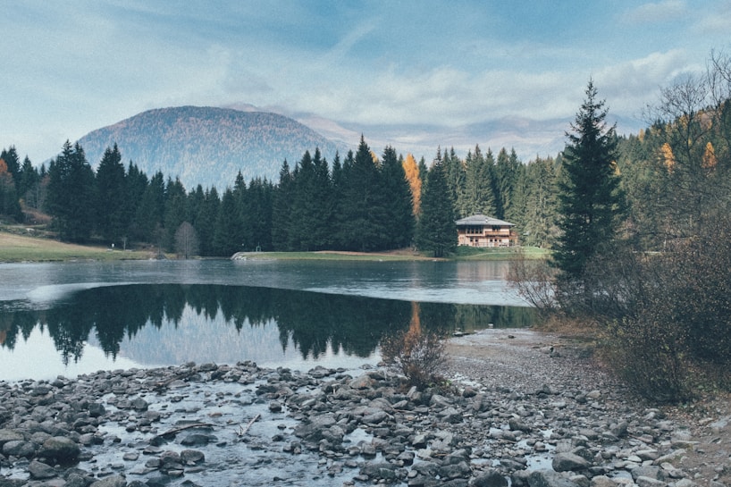 A serene landscape featuring a clear, reflective lake surrounded by dense pine forests. A small cabin can be seen nestled among the trees on the right side of the image. In the background, a mountain peak rises, partially covered by mist. The foreground contains a rocky shoreline with scattered stones, giving a natural and rustic feel.