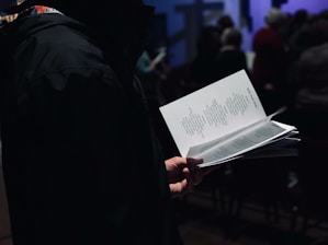 A person is holding a booklet or pamphlet with text printed on it, likely reading or following along with the content. The scene appears to be indoors with a group of people in the background, possibly at an event or gathering. The setting is dimly lit, creating a focused and intimate atmosphere.