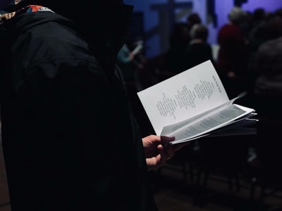 A person is holding a booklet or pamphlet with text printed on it, likely reading or following along with the content. The scene appears to be indoors with a group of people in the background, possibly at an event or gathering. The setting is dimly lit, creating a focused and intimate atmosphere.