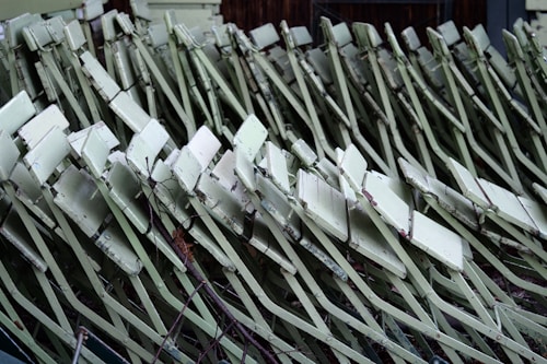 A collection of light green metal folding chairs stacked together, showing signs of age and wear with chipped paint and rust. They are arranged closely in a repeating pattern, creating an abstract geometric visual.