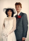 A couple is posed for a formal portrait. The woman wears a white wedding dress with lace details and a wide-brimmed hat, holding a bouquet of flowers. The man is dressed in a gray suit with a red tie and a red carnation boutonniere. Both display a calm and composed demeanor against a plain backdrop.
