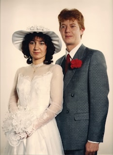A couple is posed for a formal portrait. The woman wears a white wedding dress with lace details and a wide-brimmed hat, holding a bouquet of flowers. The man is dressed in a gray suit with a red tie and a red carnation boutonniere. Both display a calm and composed demeanor against a plain backdrop.
