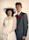 A couple is posed for a formal portrait. The woman wears a white wedding dress with lace details and a wide-brimmed hat, holding a bouquet of flowers. The man is dressed in a gray suit with a red tie and a red carnation boutonniere. Both display a calm and composed demeanor against a plain backdrop.