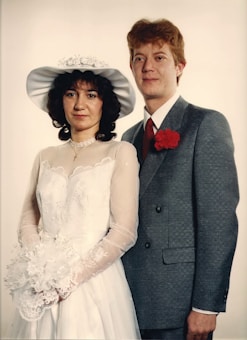 A couple is posed for a formal portrait. The woman wears a white wedding dress with lace details and a wide-brimmed hat, holding a bouquet of flowers. The man is dressed in a gray suit with a red tie and a red carnation boutonniere. Both display a calm and composed demeanor against a plain backdrop.