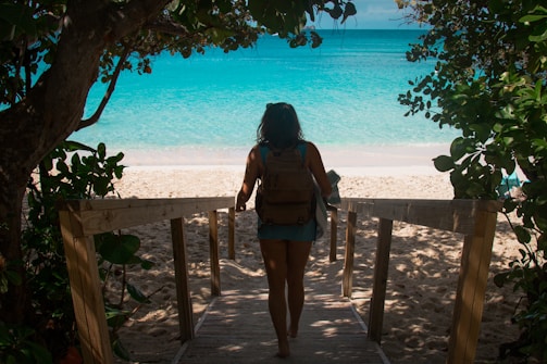 A person carrying a backpack walks down a wooden pathway leading to a sandy beach with vibrant turquoise waters. Lush green foliage surrounds the path, creating a natural frame for the scene.