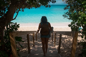 A person carrying a backpack walks down a wooden pathway leading to a sandy beach with vibrant turquoise waters. Lush green foliage surrounds the path, creating a natural frame for the scene.