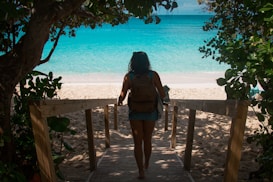 A person carrying a backpack walks down a wooden pathway leading to a sandy beach with vibrant turquoise waters. Lush green foliage surrounds the path, creating a natural frame for the scene.