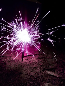 A bright, vibrant sparkler emits light and sparks, illuminating the surrounding area with a vivid pink and white glow. The sparkling light creates an energetic display against a dark background, adding a dynamic effect.