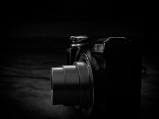 A digital camera with its lens extended, photographed with a low-key lighting effect, creating strong shadows and highlights.