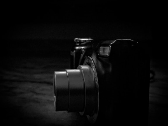 A digital camera with its lens extended, photographed with a low-key lighting effect, creating strong shadows and highlights.