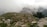 A lush green hillside with scattered rocks and a small stone hut with a metal roof in the foreground. Several colorful tents are pitched along the hilltop, surrounded by fog and low clouds. Dense trees and a cow are visible, creating a tranquil and natural setting.