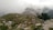 A lush green hillside with scattered rocks and a small stone hut with a metal roof in the foreground. Several colorful tents are pitched along the hilltop, surrounded by fog and low clouds. Dense trees and a cow are visible, creating a tranquil and natural setting.