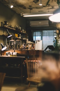 A cozy cafe interior with dim lighting. Wooden shelves lined with glass jars and coffee equipment adorn the wall. A counter displays coffee-related items and a menu, with a table in the foreground featuring a plaid tablecloth. The ambiance is warm and slightly rustic, enhanced by visible light fixtures and an air conditioning unit.