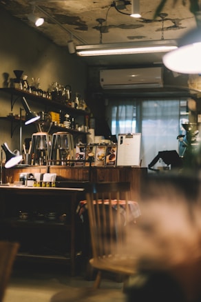 A cozy cafe interior with dim lighting. Wooden shelves lined with glass jars and coffee equipment adorn the wall. A counter displays coffee-related items and a menu, with a table in the foreground featuring a plaid tablecloth. The ambiance is warm and slightly rustic, enhanced by visible light fixtures and an air conditioning unit.