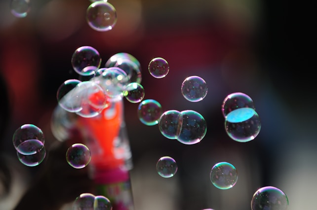 Bubbles of varying sizes float in the air, reflecting light and showing iridescent colors. In the background, a blurred and colorful object appears, possibly a bubble wand or toy.
