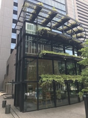 A modern glass building with metal framing and multiple levels. The structure features greenery and plants integrated into the design, including on terraces and a canopy. The lower floor has a few posters and a big glass entrance.