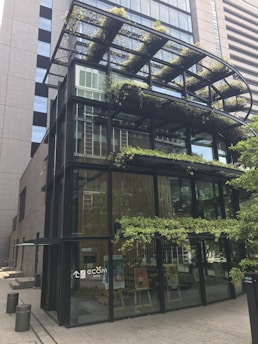 A modern glass building with metal framing and multiple levels. The structure features greenery and plants integrated into the design, including on terraces and a canopy. The lower floor has a few posters and a big glass entrance.