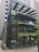 A modern glass building with metal framing and multiple levels. The structure features greenery and plants integrated into the design, including on terraces and a canopy. The lower floor has a few posters and a big glass entrance.