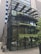 A modern glass building with metal framing and multiple levels. The structure features greenery and plants integrated into the design, including on terraces and a canopy. The lower floor has a few posters and a big glass entrance.