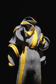 A highly stylized figure dressed in an intricate outfit composed of black and white vertical stripes interwoven with bold, yellow curved elements. The design appears avant-garde with exaggerated, voluminous spirals emerging from the outfit, contrasted starkly against a deep black background.