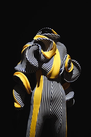 A highly stylized figure dressed in an intricate outfit composed of black and white vertical stripes interwoven with bold, yellow curved elements. The design appears avant-garde with exaggerated, voluminous spirals emerging from the outfit, contrasted starkly against a deep black background.