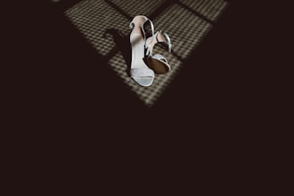 A pair of elegant high-heeled shoes are placed on a textured floor, partially illuminated by a pattern of light and shadows coming from a nearby window.