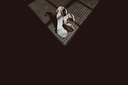 A pair of elegant high-heeled shoes are placed on a textured floor, partially illuminated by a pattern of light and shadows coming from a nearby window.
