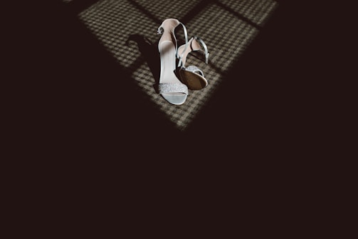 A pair of elegant high-heeled shoes are placed on a textured floor, partially illuminated by a pattern of light and shadows coming from a nearby window.