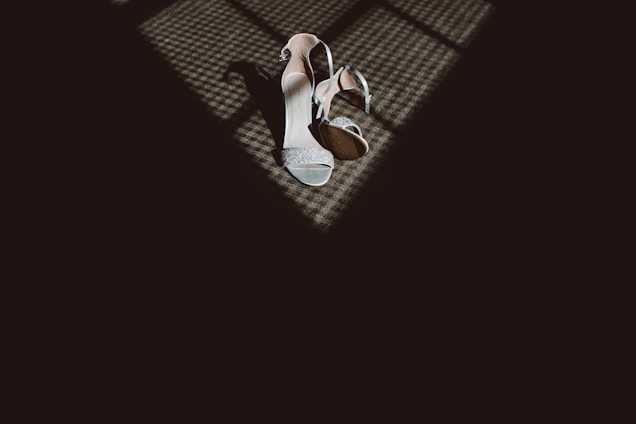 A pair of elegant high-heeled shoes are placed on a textured floor, partially illuminated by a pattern of light and shadows coming from a nearby window.