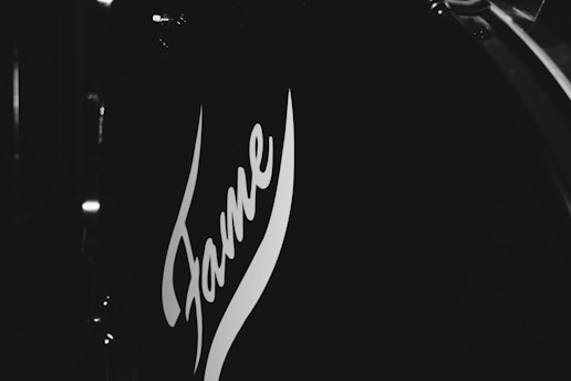 A close-up view of a drum with the word 'Fame' in stylized lettering on its surface. The image is in black and white, emphasizing the contrast between the dark drum shell and the white lettering.