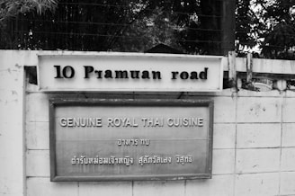 A black and white photograph of a sign attached to a wall. The sign reads '10 Pramuan road' at the top, followed by a second sign below that reads 'GENUINE ROYAL THAI CUISINE' with additional text in Thai script. The wall is made of concrete, and there are trees and some fencing visible in the background.