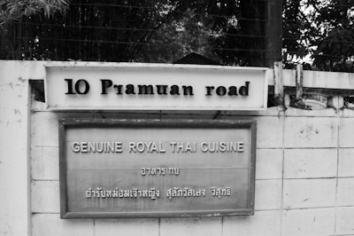 A black and white photograph of a sign attached to a wall. The sign reads '10 Pramuan road' at the top, followed by a second sign below that reads 'GENUINE ROYAL THAI CUISINE' with additional text in Thai script. The wall is made of concrete, and there are trees and some fencing visible in the background.