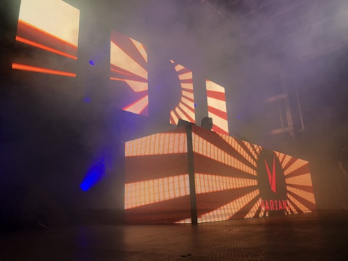 A dimly lit stage with an illuminated DJ booth featuring vibrant, geometric patterns on large LED screens. The atmosphere appears hazy with smoke, enhancing the concert-like setting. A figure, possibly a DJ, is barely visible behind the booth.