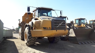 A large yellow construction vehicle, likely a dump truck, is stationed on a dirt surface. The vehicle is flanked by other construction machinery in the background, including a loader with a large bucket. The setting appears to be an industrial or construction site with equipment arranged in neat rows.