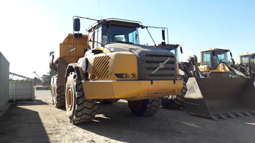 A large yellow construction vehicle, likely a dump truck, is stationed on a dirt surface. The vehicle is flanked by other construction machinery in the background, including a loader with a large bucket. The setting appears to be an industrial or construction site with equipment arranged in neat rows.