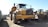 A large yellow construction vehicle, likely a dump truck, is stationed on a dirt surface. The vehicle is flanked by other construction machinery in the background, including a loader with a large bucket. The setting appears to be an industrial or construction site with equipment arranged in neat rows.