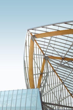 A modern architectural structure featuring a large wooden beam-supported framework with glass panels. The design showcases an intricate geometric pattern with metal supports and a clean, minimalistic aesthetic against a clear sky backdrop.