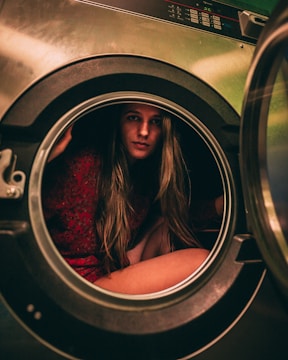 A person is sitting inside a washing machine, looking directly at the camera. The interior and exterior of the washing machine are visible, along with control panels. The person has long hair and is wearing a red patterned outfit.