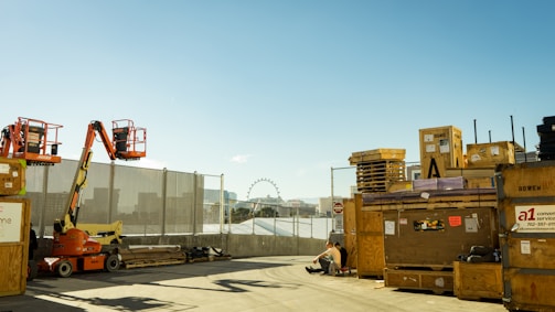 Construction equipment and pallets are stacked near a chain-link fence, with city buildings and a Ferris wheel visible in the distance under a clear blue sky. A person is sitting on the ground near the crates.