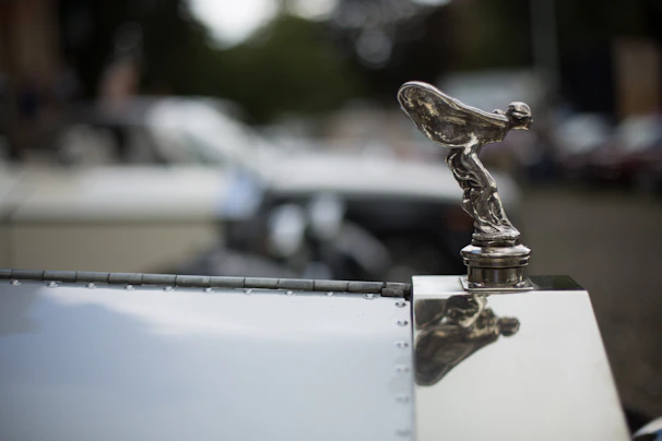 Close-up of a gleaming hood ornament reflecting the sunset at a lively car meet hosted by Athena Car Collective.