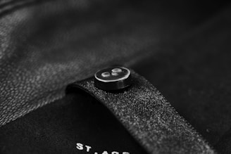A close-up of a black button sewn onto a black piece of fabric, possibly a garment. The surface has a slightly reflective texture, and there is subtle lighting creating soft highlights.