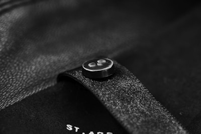 A close-up of a black button sewn onto a black piece of fabric, possibly a garment. The surface has a slightly reflective texture, and there is subtle lighting creating soft highlights.