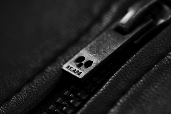 Close-up view of a metallic zipper slider against a textured, dark fabric background. The word 'SF-ARE' is engraved on the slider, and the photograph is captured in black and white, emphasizing the details of the metal and fabric.