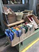 A display of colorful candles arranged neatly in a wooden holder is in the foreground. Above, woven baskets and a crate containing other items are placed on a wooden shelf. The setting appears to be a shop with a window displaying the text 'Brixton cornercopia'.