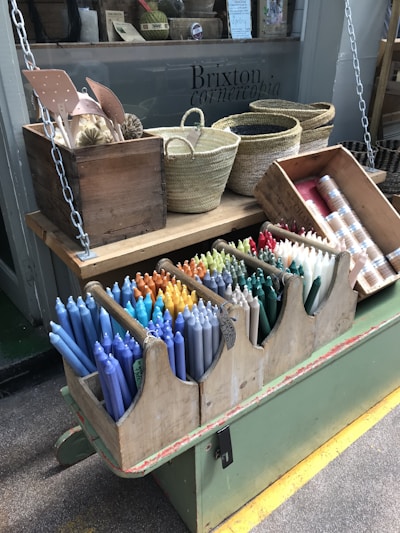 A display of colorful candles arranged neatly in a wooden holder is in the foreground. Above, woven baskets and a crate containing other items are placed on a wooden shelf. The setting appears to be a shop with a window displaying the text 'Brixton cornercopia'.