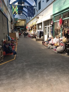 A covered market corridor with various shops on either side. The left side has a closed shop with chairs and colorful cushions displayed outside. The right side features shops showcasing clothes, bags, and textiles. Several flags hang from the ceiling, and a few people are walking further down the corridor.