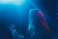 A person with curly hair is playing a guitar under blue and red stage lights, creating a moody and atmospheric scene. The focus on the individual and instrument suggests a live music performance.