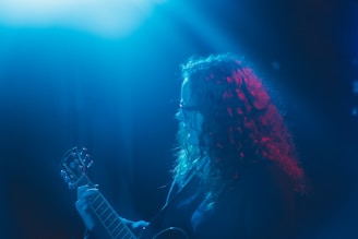 A person with curly hair is playing a guitar under blue and red stage lights, creating a moody and atmospheric scene. The focus on the individual and instrument suggests a live music performance.