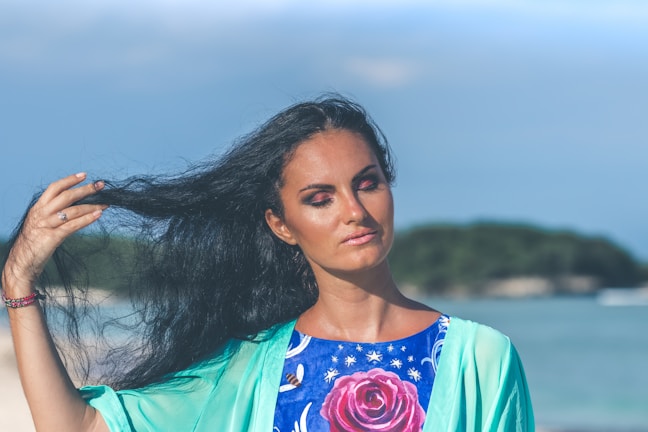 A woman with long dark hair is standing near a beach, gently holding a strand of her hair. She wears a brightly colored top featuring a large pink rose and a light turquoise cover-up. The background shows a blurred seascape with a green island in the distance under a clear blue sky.