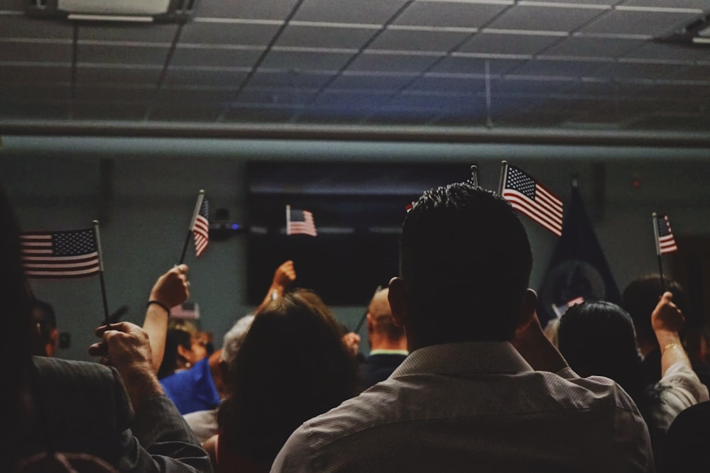 A crowd of seated people of various races and genders face away from the camera & towards a dark stage. They all hold up miniature American flags.